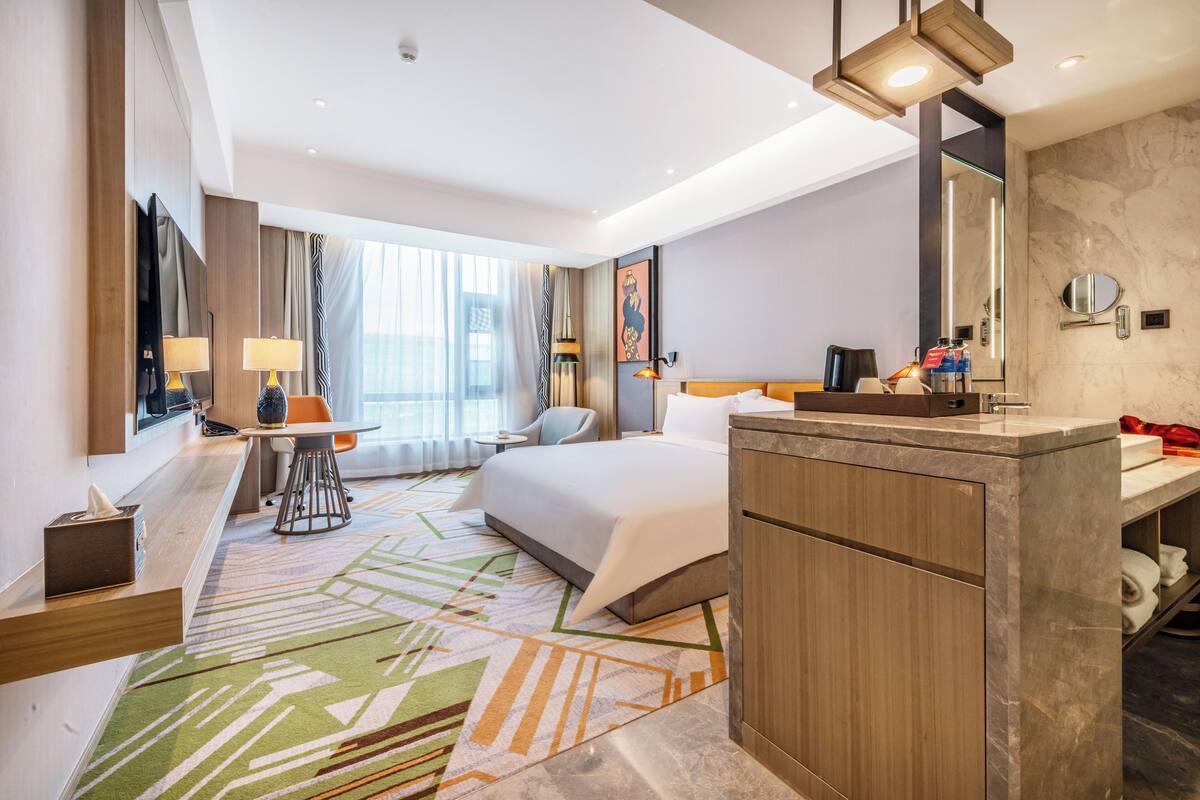 Une chambre d’hôtel avec un grand lit, un bureau avec une chaise, une salle de bain avec un miroir et un lavabo, et un tapis aux couleurs vives.