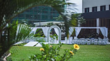 An outdoor wedding setup with white chairs and arches, a grassy area, and a building in the background.