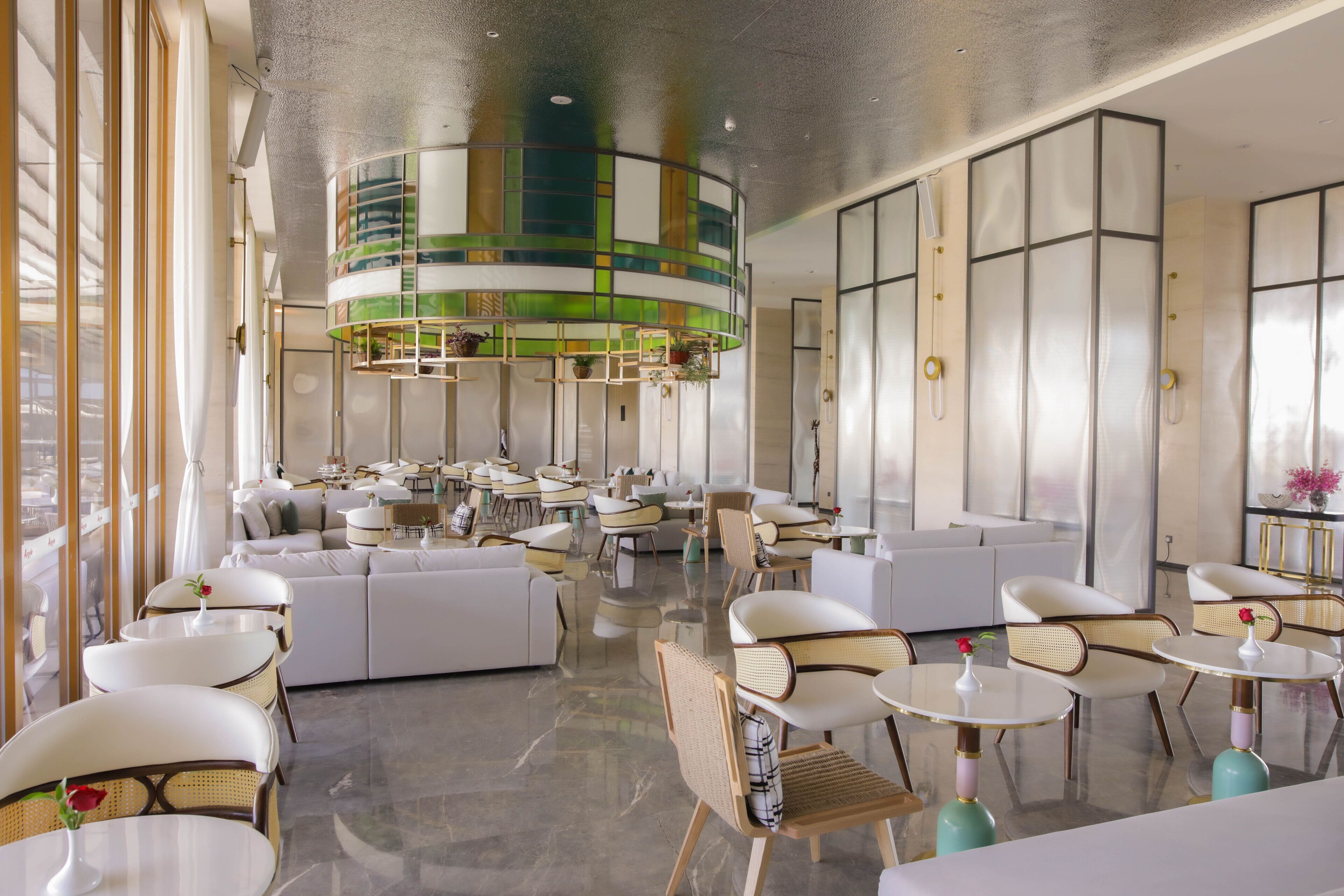 A modern hotel lobby with a central seating area, multiple tables, and a distinctive green and white patterned ceiling.