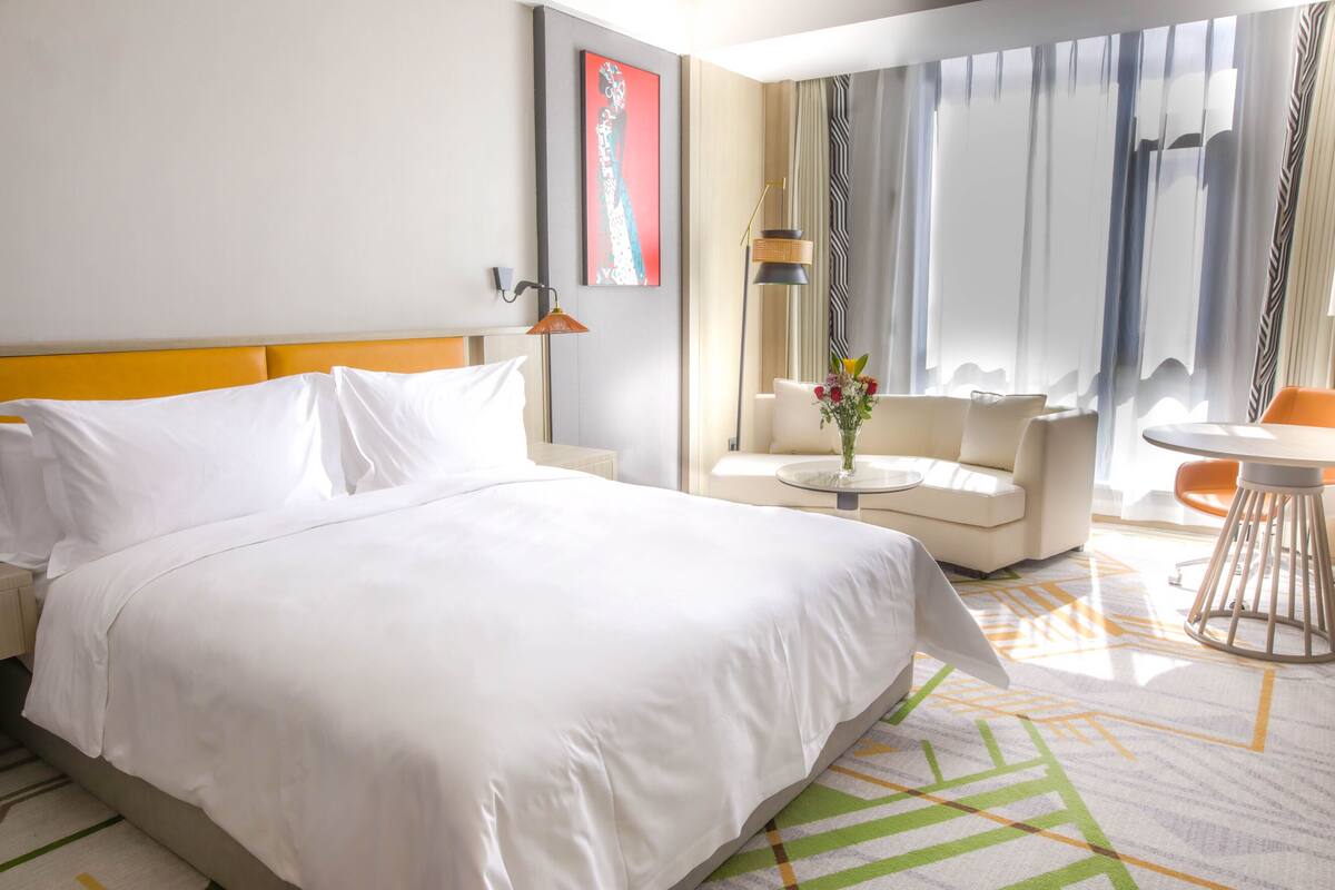 Une chambre d’hôtel avec un grand lit, un canapé, une petite table ronde ornée d’un vase, et une vue sur l’extérieur grâce à de grandes fenêtres.