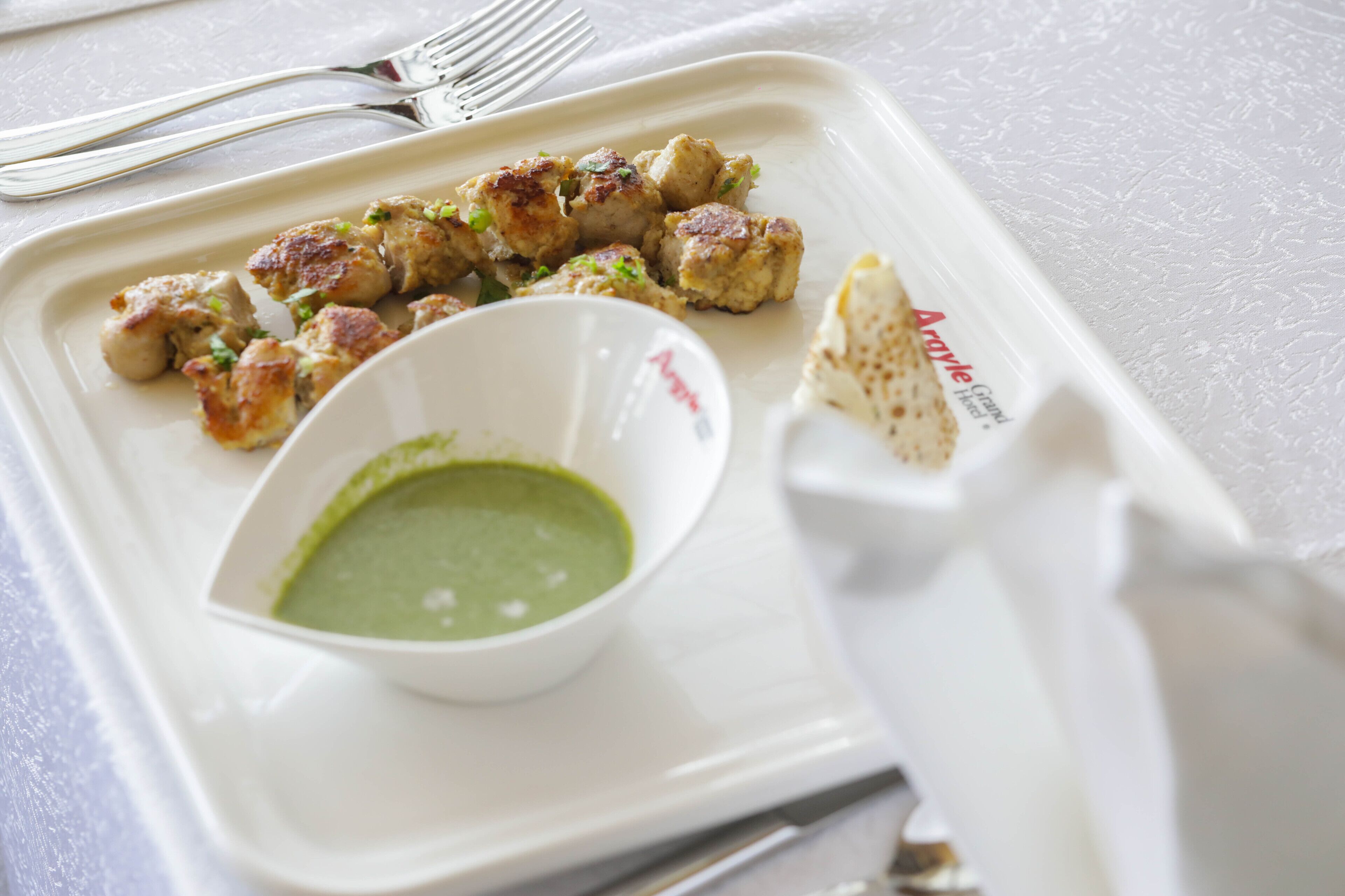 Une assiette blanche contenant des brochettes de poulet grillé et un bol de sauce verte, accompagnées d’une serviette pliée et d’une fourchette, le tout posé sur une table.