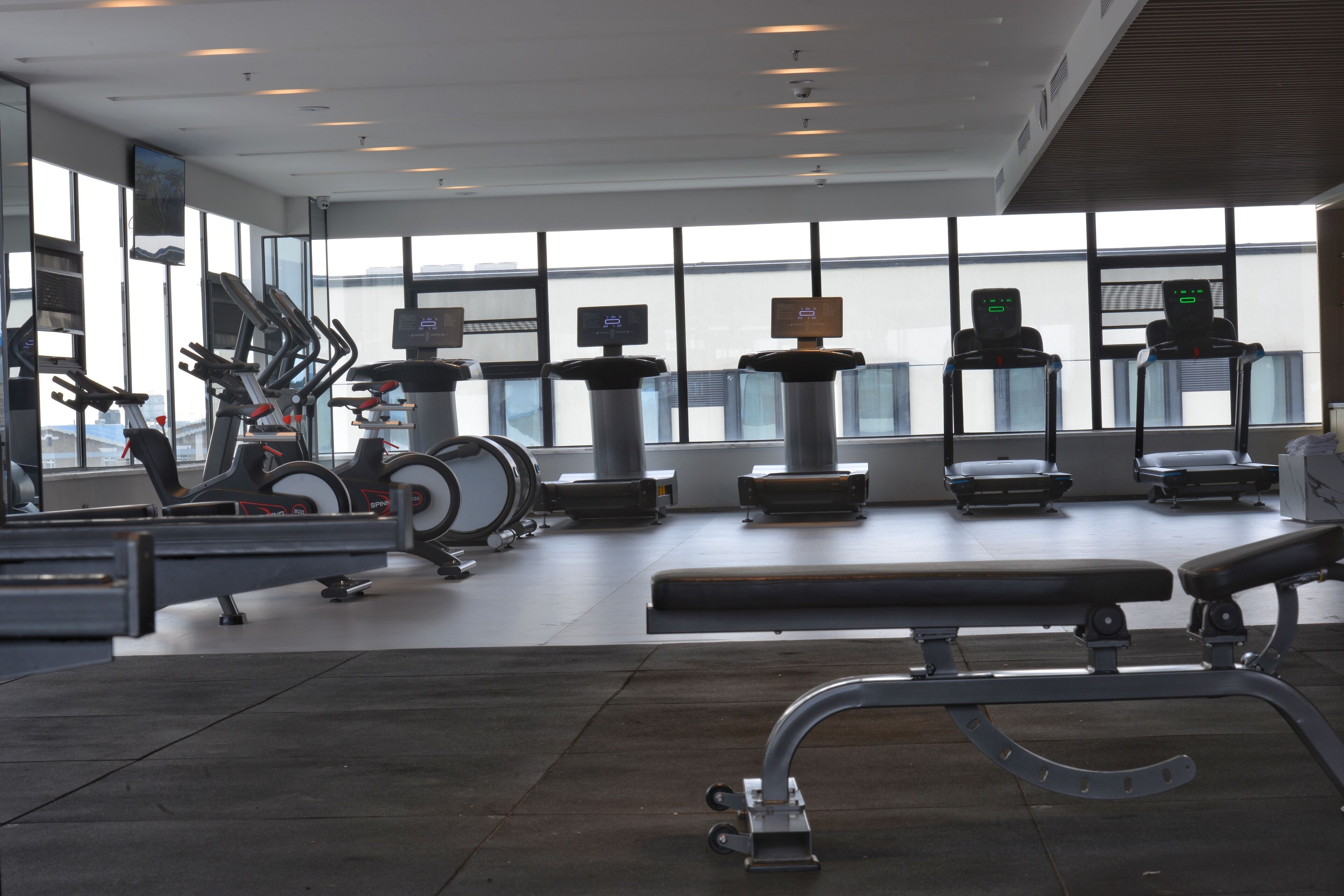Un gimnasio con equipos de ejercicio variados, ventanas amplias y un banco.