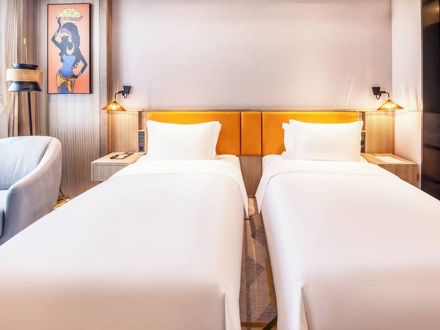 A hotel room with two single beds, a chair, a nightstand with a lamp, and a wall-mounted artwork.