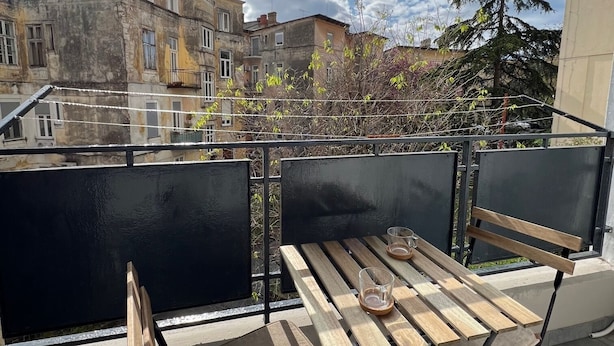 Ein Balkon mit Holztisch und -stühlen, der auf eine Stadtansicht mit historischen Gebäuden und blauem Himmel blickt.