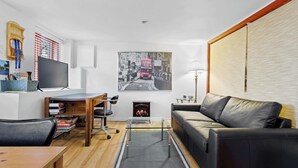 A modern living room with a black leather sofa, a glass coffee table, a wooden desk, a flat-screen TV, a fireplace, and a large cityscape painting on the wall. - Free ferry | Aux Trois Filles (Lévis)