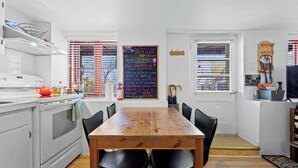 A modern kitchen with white appliances, a wooden dining table, and black chairs. There is a chalkboard with messages, a window with blinds, and a small shelf with items. - Free ferry | Aux Trois Filles (Lévis)
