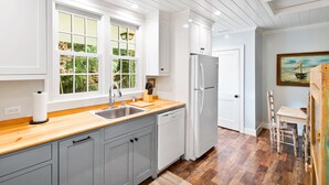 Una cocina con armarios blancos, encimera de madera, refrigerador blanco y ventana con vistas a zonas verdes.