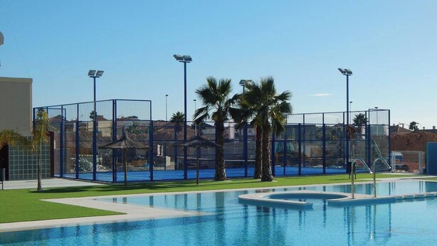 A swimming pool with a clear blue water, surrounded by a fenced area with palm trees and a building in the background. - Groundfloor Varudi Aguas Nuevas, Torrevieja (Torrevieja)