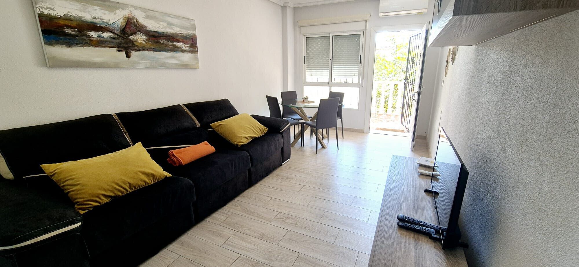 Una sala de estar con un sofá negro, cojines amarillos y anaranjados, una mesa de centro de vidrio y un cuadro en la pared.