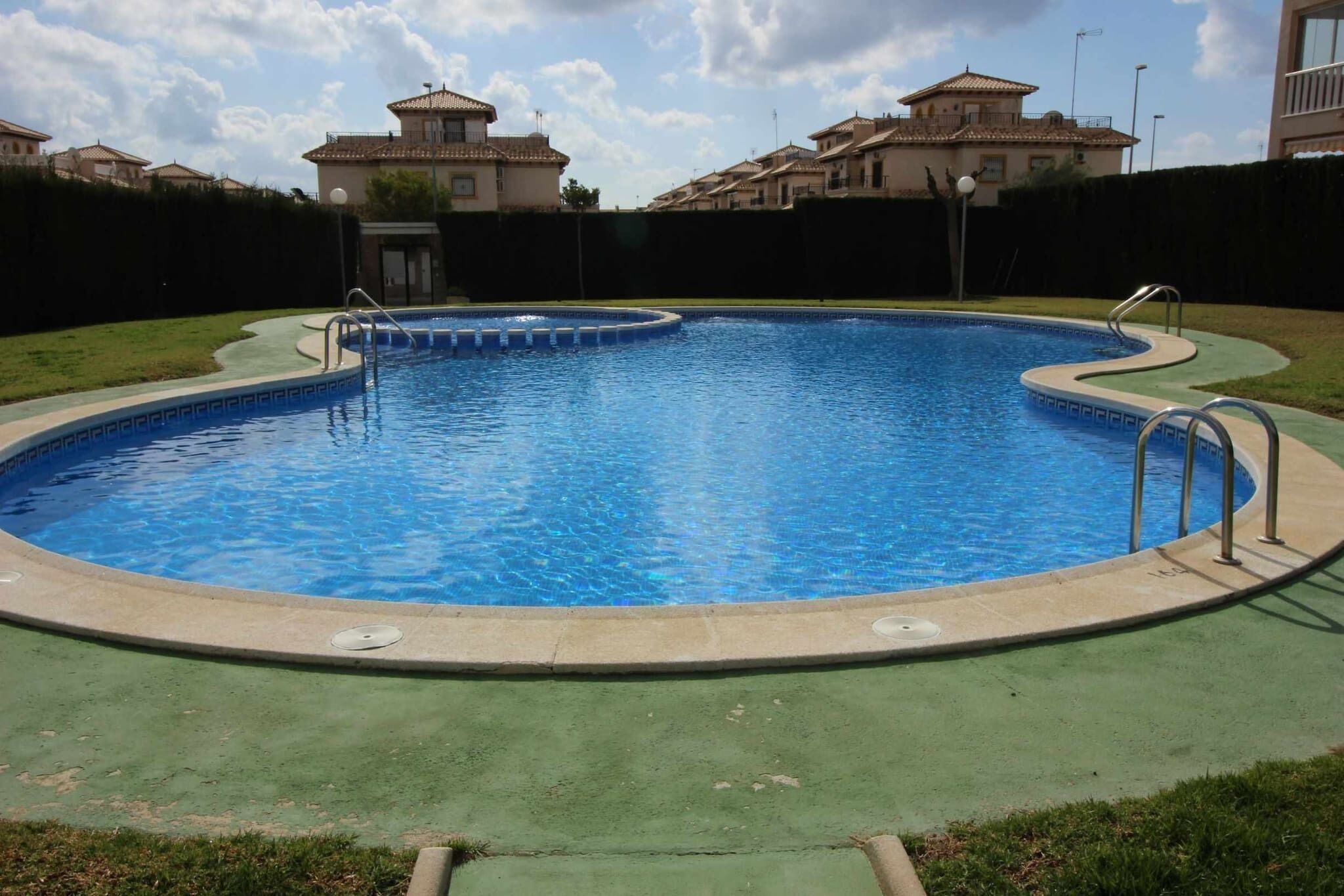 En swimmingpool med ett grönt poolområde, omgiven av en häck och bostadshus i bakgrunden.