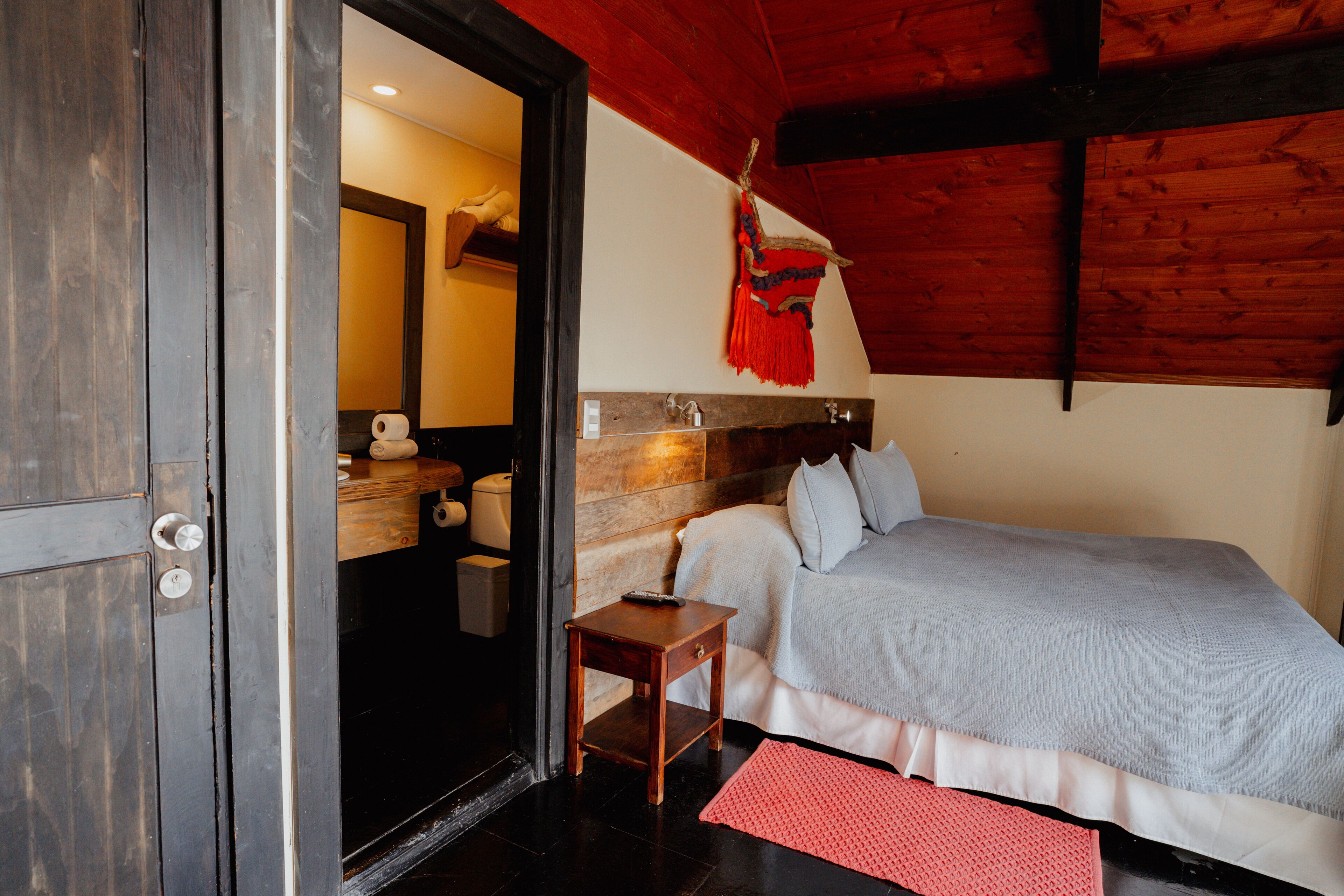 A bedroom with a bed, a wooden headboard, a small wooden nightstand, a red rug, and a bathroom visible through an open door.