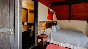 A bedroom with a bed, a wooden headboard, a small wooden nightstand, a red rug, and a bathroom visible through an open door.