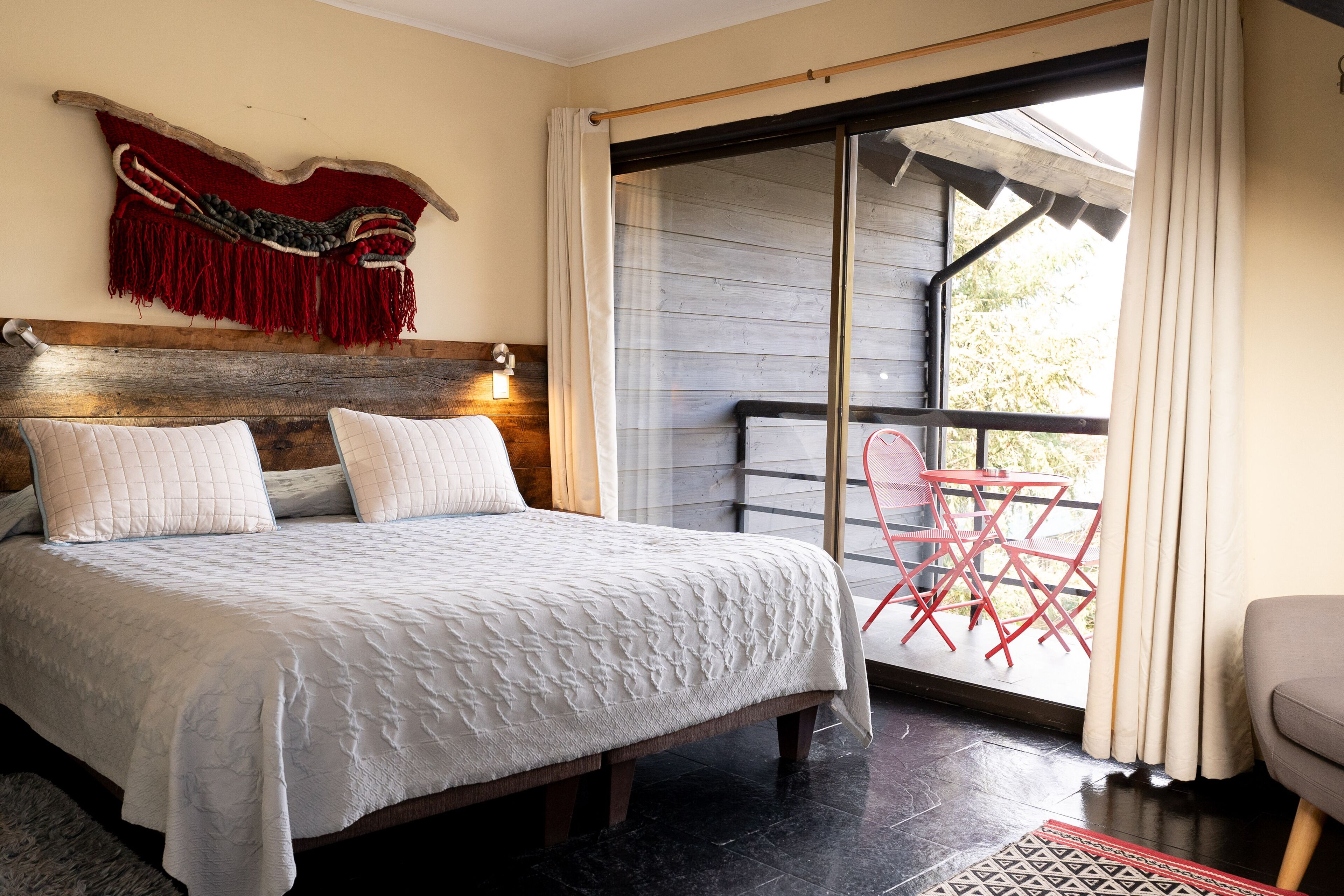 A bedroom with a large bed, a wooden headboard, a red and white wall hanging, and a balcony with red chairs.