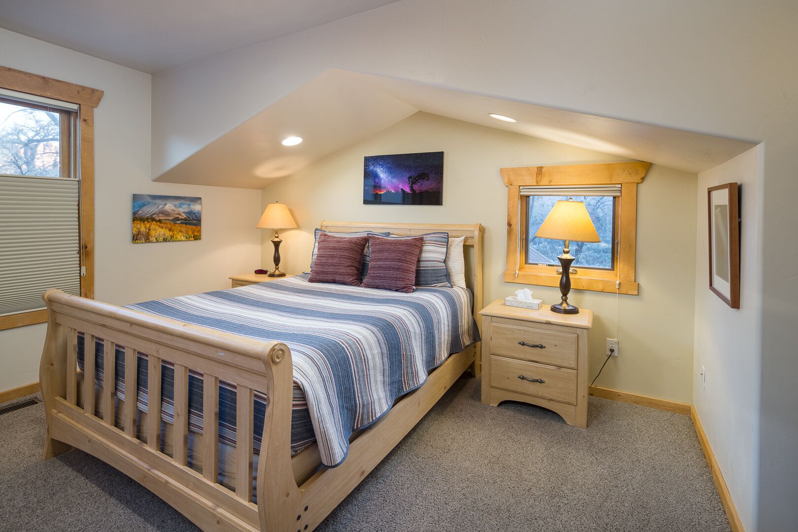 A bedroom with a wooden bed, a nightstand, a lamp, a window with a view, and a painting on the wall.