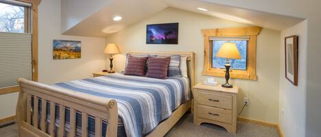 A bedroom with a wooden bed, a nightstand, a lamp, a window with a view, and a painting on the wall.