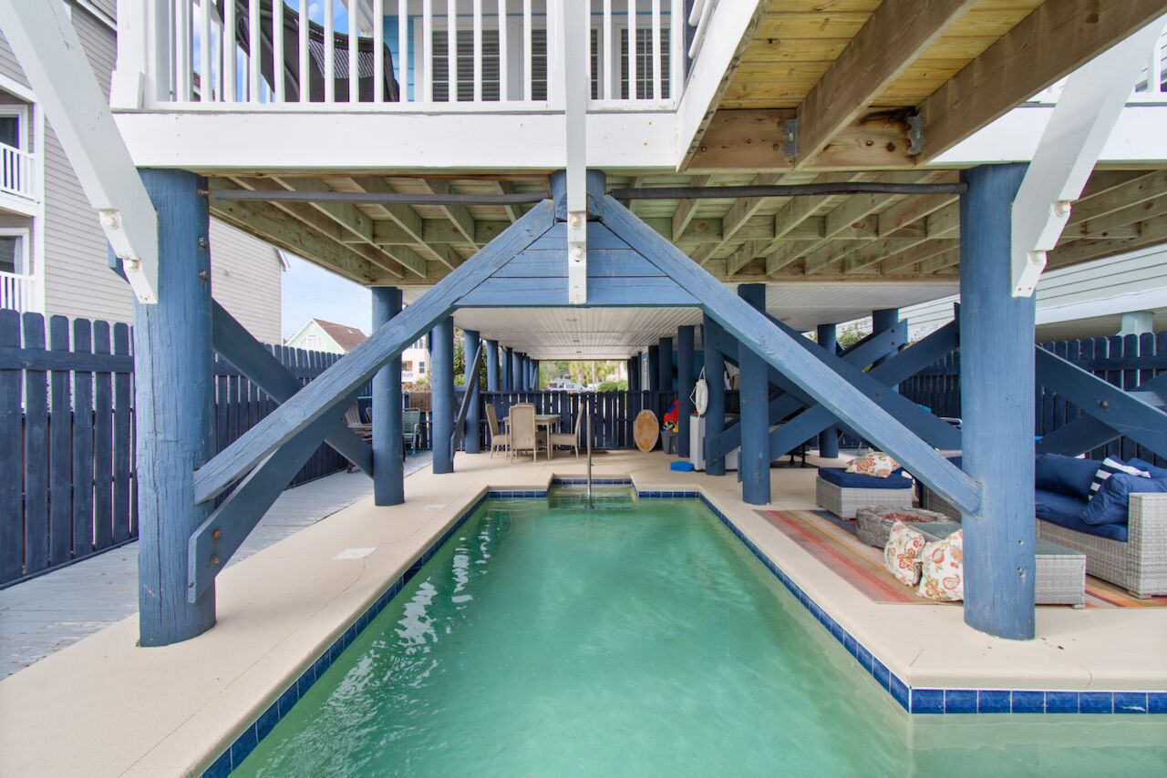 A covered swimming pool with a wooden deck and blue pillars, surrounded by a fence and outdoor furniture.