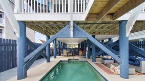 A covered swimming pool with a wooden deck and blue pillars, surrounded by a fence and outdoor furniture.