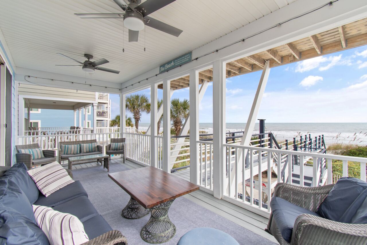 A covered outdoor area with a wooden table, sofas, and a view of the beach.