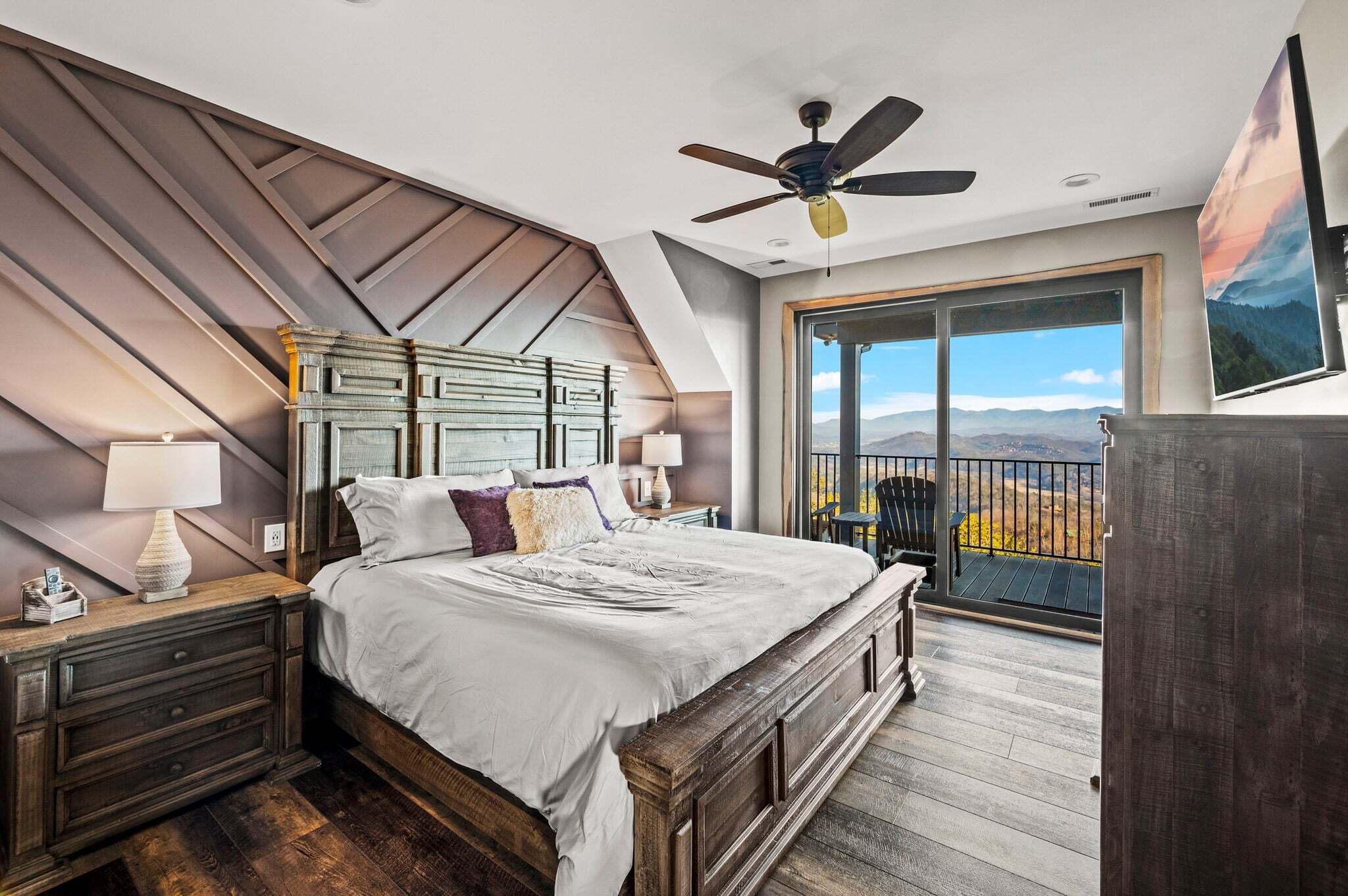 A bedroom with a large bed, a ceiling fan, a nightstand, a dresser, and a balcony with a view of the mountains.