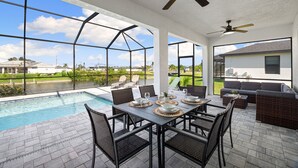 A poolside dining area with a table set for six, surrounded by wicker chairs, overlooking a swimming pool and a view of a residential area. - New build from 2023 impresses with its modern yet cozy interior and heated pool (Cape Coral)