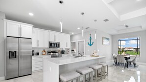 A modern kitchen with white cabinetry, stainless steel appliances, and a large island with bar stools. - New build from 2023 impresses with its modern yet cozy interior and heated pool (Cape Coral)