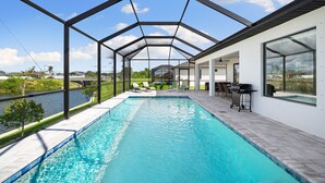 A modern outdoor pool with a glass enclosure, a patio area with outdoor furniture, and a view of a river. - New build from 2023 impresses with its modern yet cozy interior and heated pool (Cape Coral)