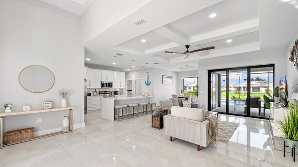 A spacious living area with a large sofa, a coffee table, and a dining area with bar stools. There is a kitchen with white cabinets and a central island. The room has a large sliding glass door leading to a patio with outdoor furniture. - New build from 2023 impresses with its modern yet cozy interior and heated pool (Cape Coral)