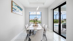 A modern dining area with a glass table, six chairs, and a large window offering a view of a pool and outdoor furniture. - New build from 2023 impresses with its modern yet cozy interior and heated pool (Cape Coral)