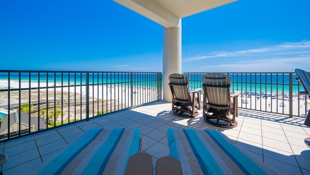 Un balcon avec deux chaises et une table, donnant sur une plage sous un ciel bleu dégagé.