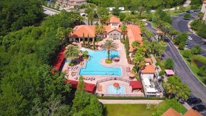 Vista aérea de un resort con piscina central, rodeado de palmeras y un edificio de techo rojo.