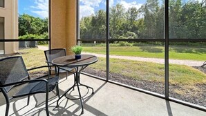 Un balcón con mesa y sillas, ventanas amplias que dan a un área verde y árboles.
