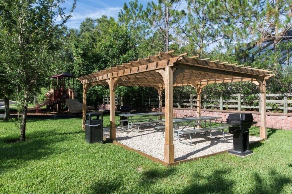 A wooden pergola with picnic tables and a barbecue grill in a park setting.