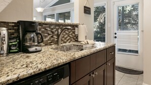 A kitchen with a granite countertop, coffee maker, dishwasher, and a window with blinds.