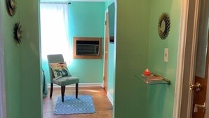 Käytävän seinät ovat turkoosin väriset, lattialla on puulattia, siellä on myös pehmustettu tuoli, pieni matto ja ovi oikealla puolella.