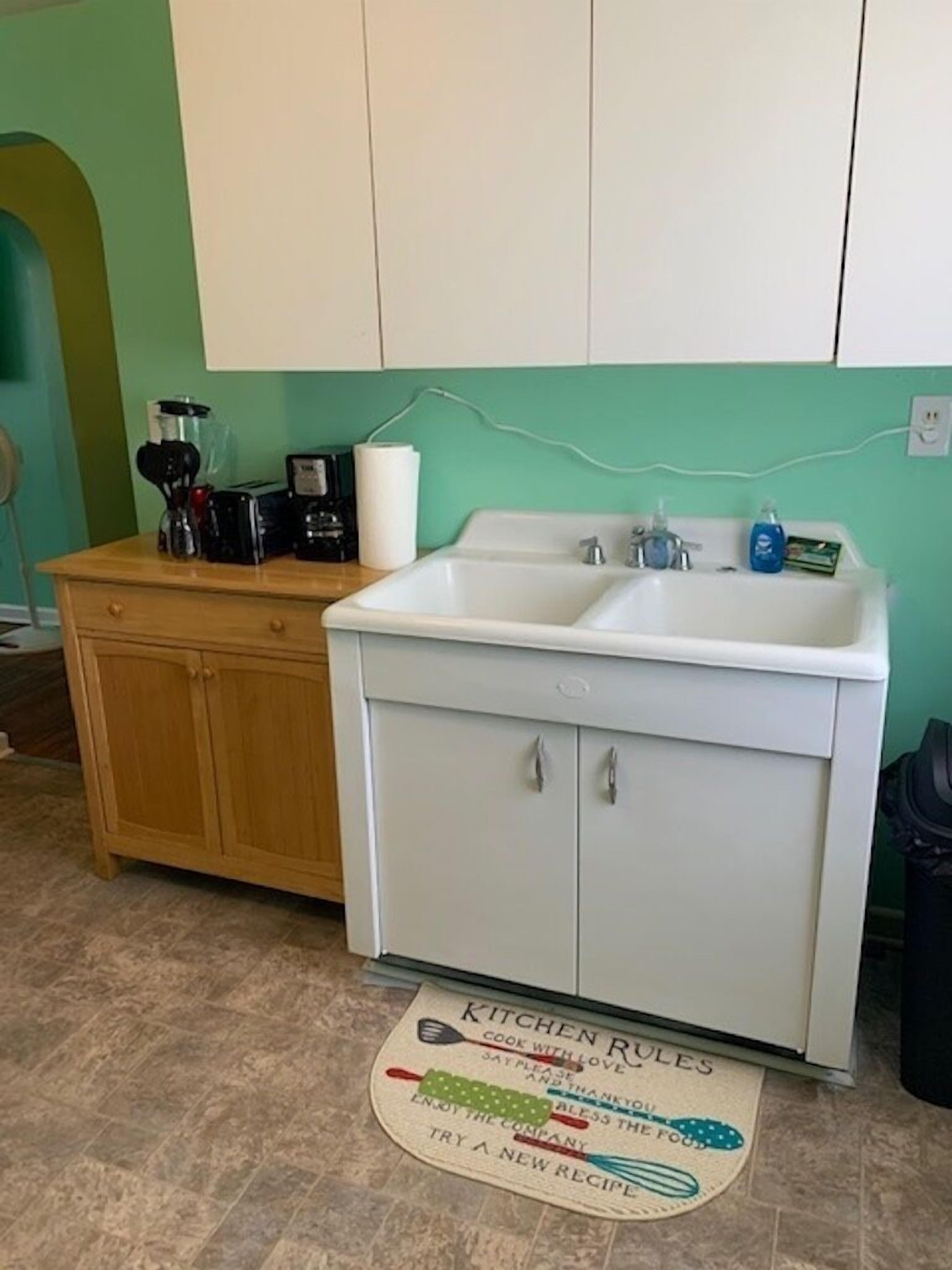 A kitchen with a white sink, wooden cabinets, and a kitchen mat with rules.