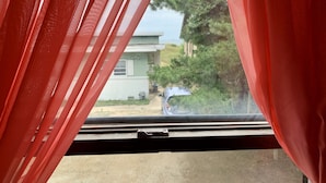 A window with red curtains partially drawn, revealing a view of a house and a parked car outside. - Break Side Studio #5 Apts by RedAwning (Norfolk)