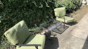 A green cushioned chair and table set on a concrete surface, surrounded by dense green foliage. - Break Side Studio #5 Apts by RedAwning (Norfolk)
