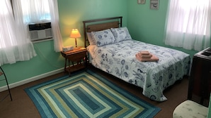 A room with a bed, a nightstand with a lamp, a radiator, and a window with curtains. - Break Side Studio #5 Apts by RedAwning (Norfolk)