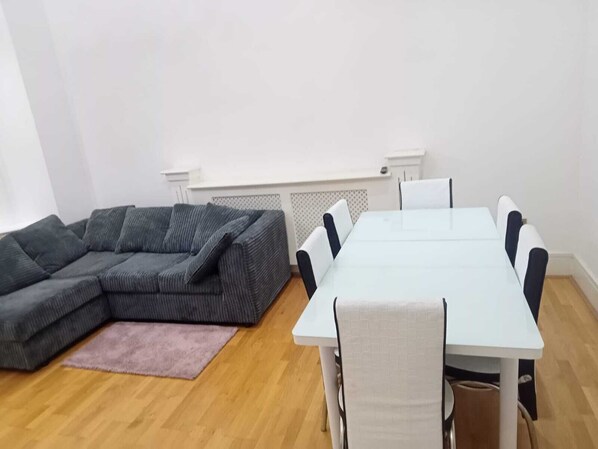 A living room with a grey sectional sofa, a white dining table with six chairs, a pink rug, and a white radiator. - Charming 2-bed Apartment in Wolverhampton (Wolverhampton)
