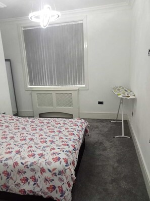 A bedroom with a bed, a window with blinds, a standing lamp, and a small table. - Charming 2-bed Apartment in Wolverhampton (Wolverhampton)