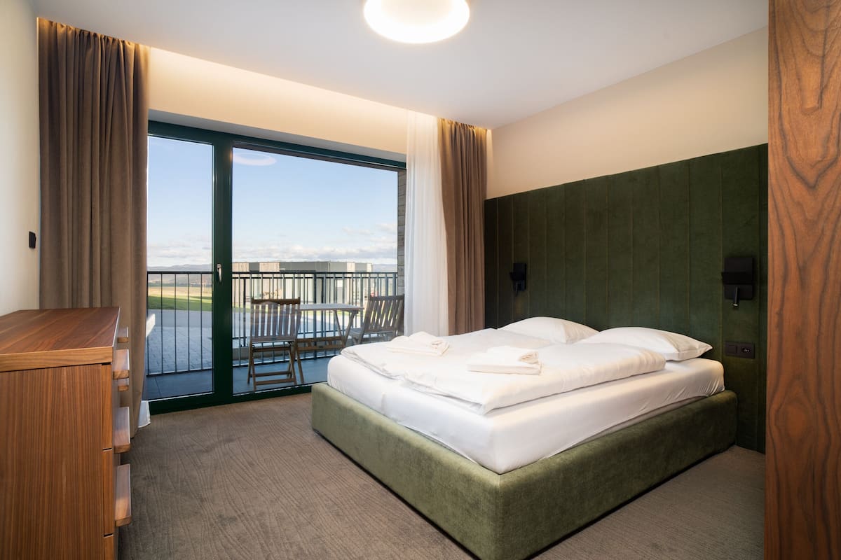 A hotel room with a bed, a wooden dresser, a balcony with a table and chairs, and a view of the city.