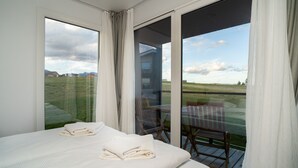 A bedroom with a large window, a bed with neatly folded white towels, and a view of a grassy field and distant buildings. - Royal Village (Malý Slavkov)