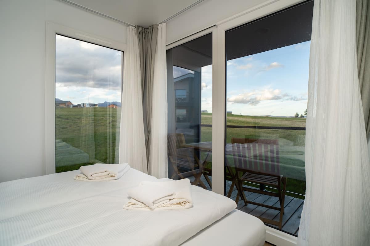 A bedroom with a large window, a bed with neatly folded white towels, and a view of a grassy field and distant buildings.