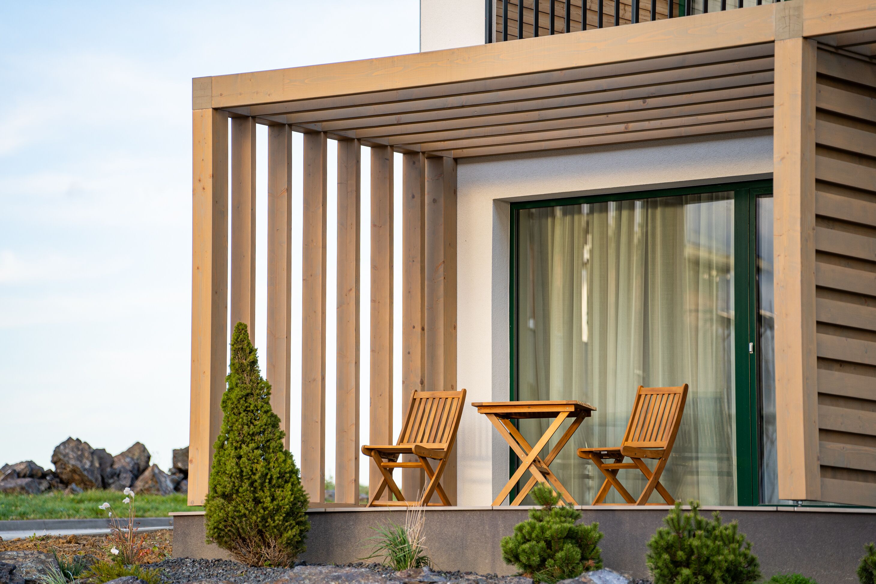 A modern building with a wooden pergola, two wooden chairs, and a table on the balcony.