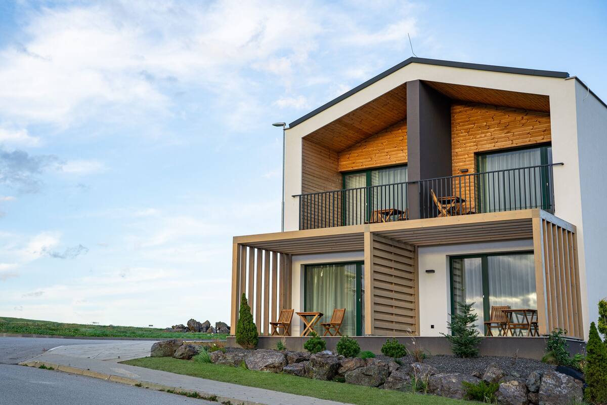 A modern two-story house with a balcony, large windows, and a stone wall in front.
