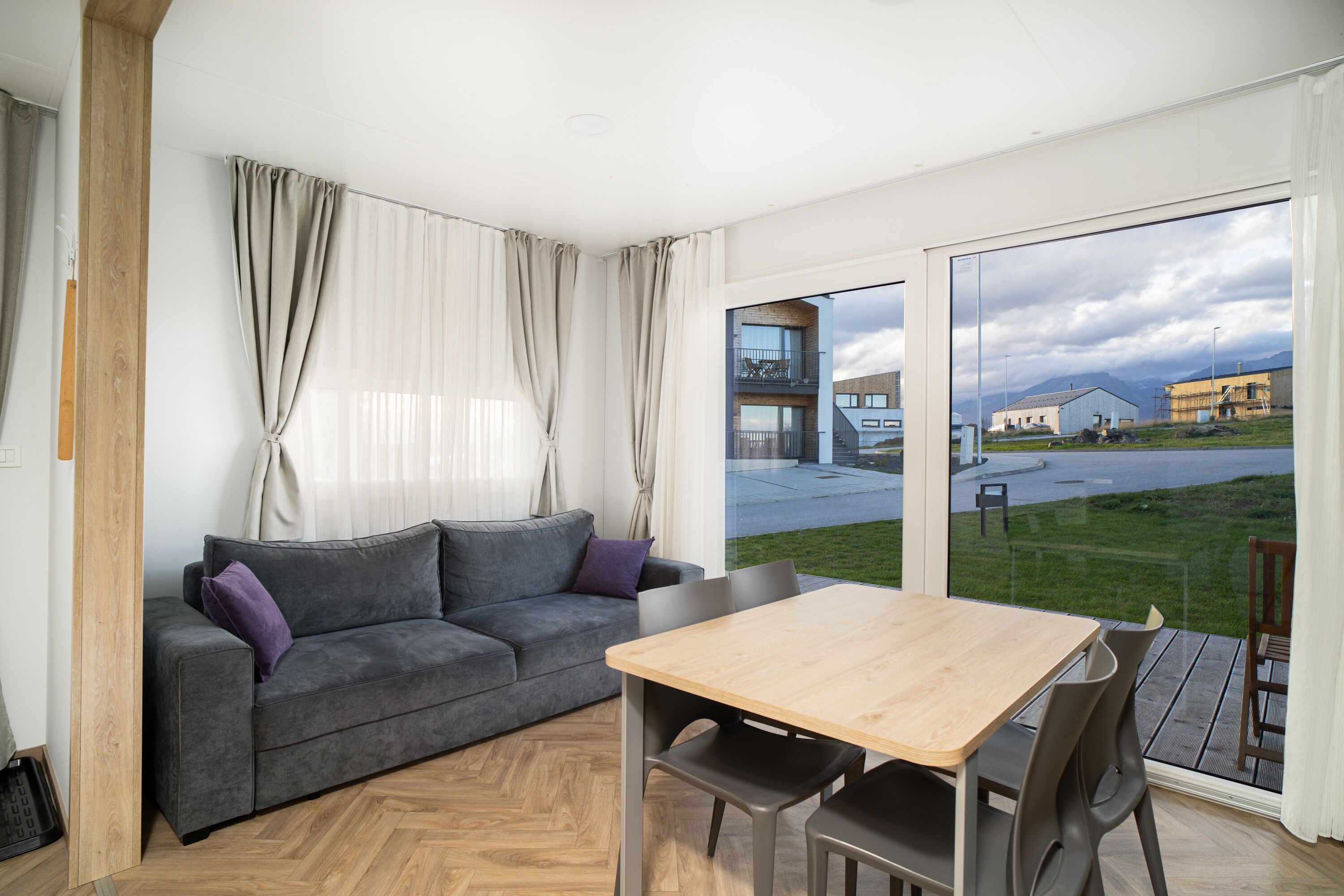 A modern living room with a grey sofa, a wooden dining table, and a view of a building and landscape through large glass doors.