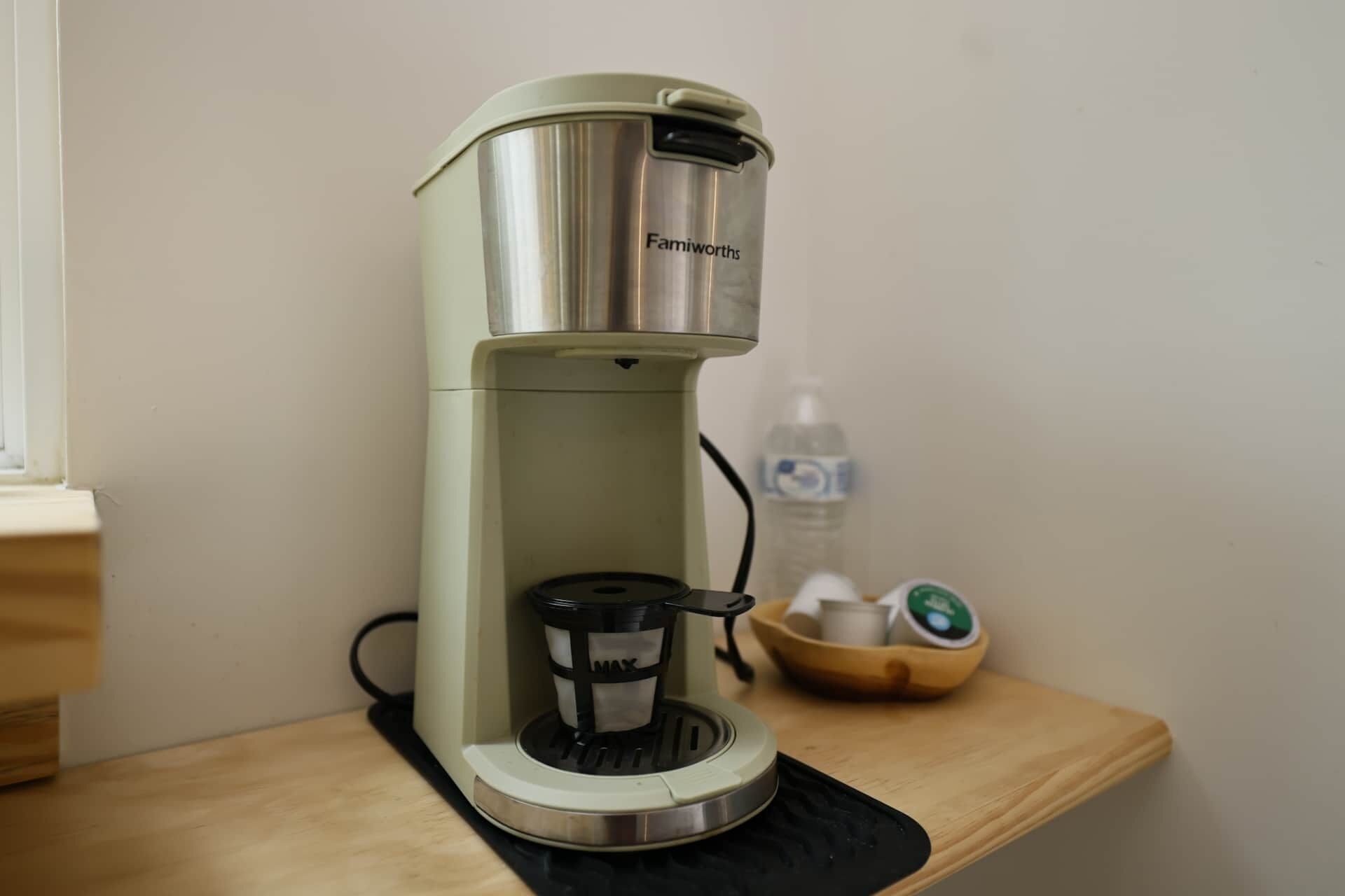 Een Famiworths koffiezetapparaat op een houten tafel, met een waterfles en een schaaltje theezakjes op de achtergrond.
