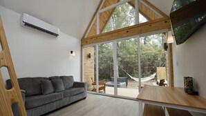A modern interior with a grey sofa, wooden beams, a large sliding glass door leading to an outdoor area with a hammock and a table. - Luxury Cabin - Riolago (Montgomery County)