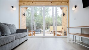 Una sala moderna con un sofá gris, piso de madera y un gran ventanal corredero que da a un espacio exterior con una hamaca y una mesa de picnic.