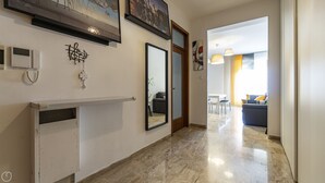 Un pasillo con piso de mármol, pared blanca con un teléfono y adornos, y una puerta que da a un área de estar con sofá y mesa.